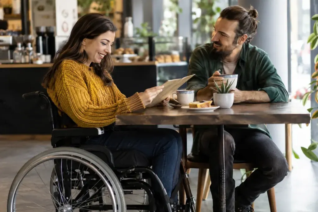 Two adults chatting at an accessible cafe table, one wheelchair user choosing a menu independently.